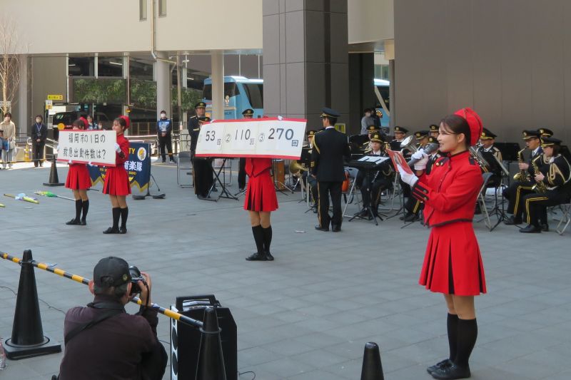 演奏をする消防音楽隊と、クイズを出題するカラーガード隊の写真