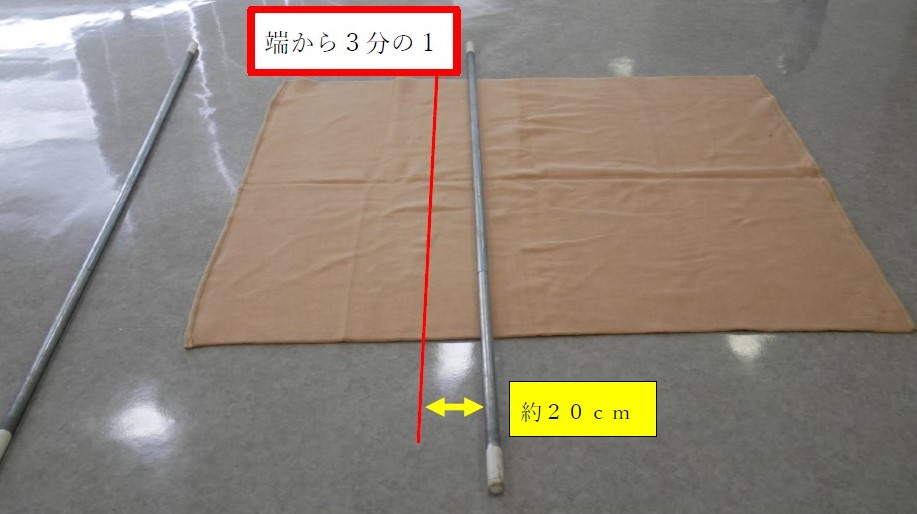 ２　床に広げた毛布の上に棒を１本置く。棒の位置は，毛布の短辺から３分の１のラインより約２０センチ毛布の内側に移動したラインで，棒の両端は毛布からはみ出るように置く。