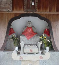 写真：中央区唐人町の成道寺境内にある八兵衛地蔵　小堂の中に祀られた数体の仏像のうち中央に鎮座する地蔵像で、石造の座像であり、赤い前掛けを掛けている