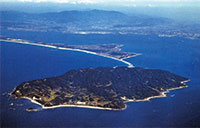 写真：志賀島を北西から南東へと俯瞰した空撮写真で、志賀島の全景、海の中道、博多湾が写し出されている。海は群青で、志賀島は緑深い