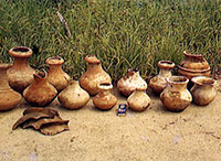 写真：板付遺跡の井戸から出土した土器。
合計13点の壺型土器とその他破片などが、前後2列で地面に並べられている。壺型土器のなかには丹塗りが施されたものがある。