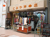 写真：川端商店街のとみや洋傘店の店頭を写したもの。オレンジ色の店名看板の下、店先にはワゴンが置かれ、傘や服飾小物などが並んでいる
