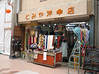 写真:川端商店街のとみや洋傘店の店頭を写したもの。オレンジ色の店名看板の下、店先にはワゴンが置かれ、傘や服飾小物などが並んでいる