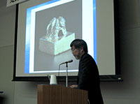 写真：演台で講演される鶴間和幸先生。背後のスクリーンには金印の写真が投影されている