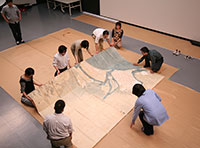 写真：九州国立博物館の撮影室で、15平方メートル以上の大型絵図を床に広げているところ。絵図は細かく折りたたまれており、7名の調査員が絵図の端を持って少しずつ広げている