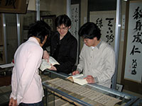 写真：玄洋社記念館で書簡の調査をしている光景。調査員が3名、ガラスケース上に置かれた古文書（書簡）を覗き込み、書かれている崩し字を解読しようと試みている