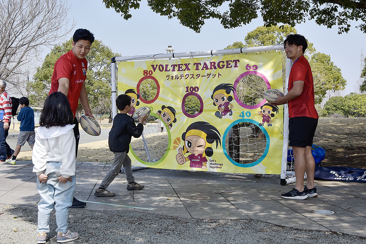プロラグビーチームであるキューデンヴォルテクスと子どもたちがラグビーボールを大中小の穴にいれていくゲーム。パスターゲットをしている写真です。